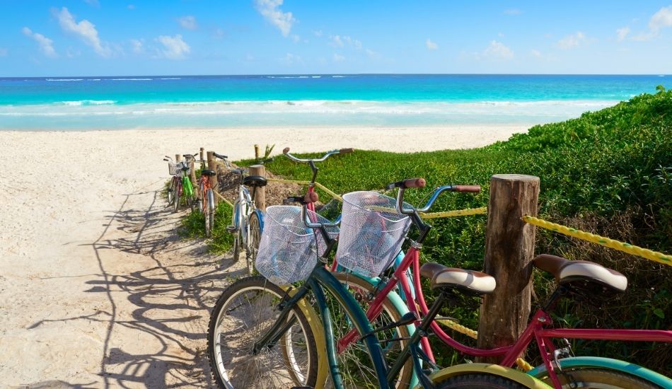 Colorful vintage bicycles parked near a sandy beach path with turquoise waters in Tulum.